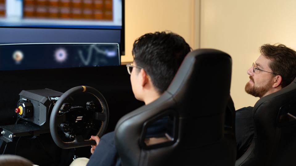 person sitting in a vehicle gaming chair holding a gaming steering wheel, looking at a screen showing a blurred prototype of the display